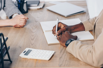 Cropped view of recruiter conducting job interview with employee and writing in notebook at table in office