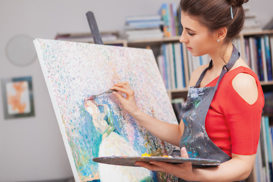 Attractive female professional artist working at her studio, painting on canvas with oil paint