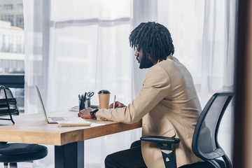 Selective focus of african american recruiter with laptop and paper cup of coffee at table in office