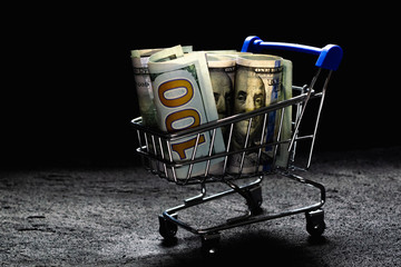 shopping cart with a blue handle with hundred dollar bills on black concrete dark background. copy...