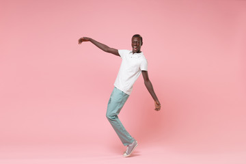 Fototapeta na wymiar Cheerful young african american man guy in white polo shirt, turquoise trousers posing isolated on pastel pink wall background. People lifestyle concept. Mock up copy space. Standing on toes, dancing.