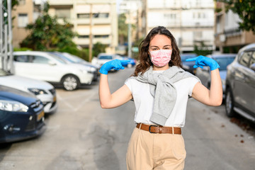 A brunette girl in protective gloves shows a medical mask that is on her face. She is standing on the street. The concept of self-defense and prevention of viruses, epidemics. Urban view. Close-up.