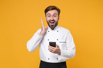 Excited young bearded male chef cook or baker man in white uniform shirt posing isolated on yellow wall background. Cooking food concept. Mock up copy space. Using mobile phone, typing sms message.