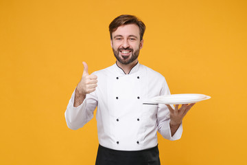 Smiling young bearded male chef cook or baker man in white uniform shirt posing isolated on yellow background in studio. Cooking food concept. Mock up copy space. Hold empty plates, showing thumb up.