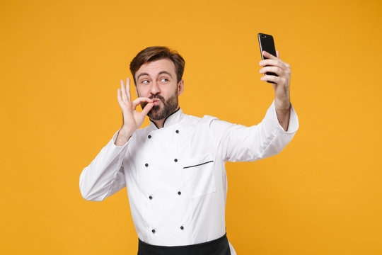 Funny bearded male chef cook or baker man in white uniform isolated on yellow background. Cooking food concept. Mock up copy space. Doing selfie shot on mobile phone, making okay taste delight sign.