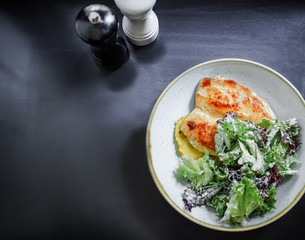 The main dish. Grilled chicken breast with a crust on a plate with mashed potatoes and green salad on a black background. Top view.