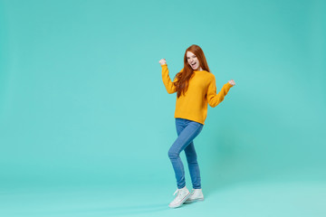 Happy young redhead woman girl in yellow sweater posing isolated on blue turquoise wall background studio portrait. People sincere emotions lifestyle concept. Mock up copy space. Doing winner gesture.