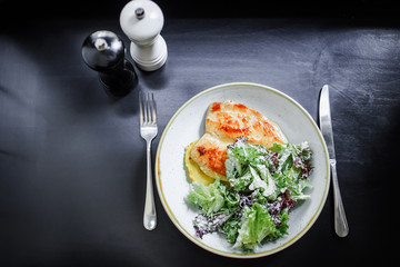 The main dish. Grilled chicken breast with a crust on a plate with mashed potatoes and green salad on a black background. Top view.