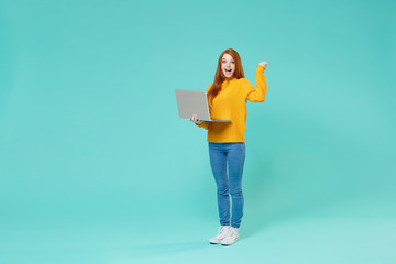 Excited young redhead girl in yellow sweater posing isolated on blue turquoise wall background in studio. People lifestyle concept. Mock up copy space. Work on laptop pc computer doing winner gesture.