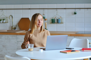 Lovely blonde woman working with her laptop while sitting in the kitchen in her apartment
