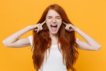 Crazy young redhead woman girl in white blank empty t-shirt posing isolated on yellow background. People lifestyle concept. Mock up copy space. Covering ears with fingers, keeping eyes closed, scream.