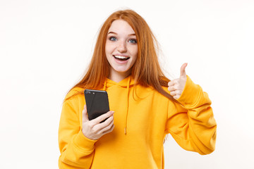Cheerful young redhead woman girl in yellow hoodie posing isolated on white background studio portrait. People emotions lifestyle concept. Mock up copy space. Using mobile phone, showing thumb up.