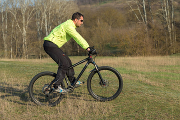 Obraz premium Cyclist in pants and green jacket on a modern carbon hardtail bike with an air suspension fork. The guy on the top of the hill rides a bike.