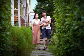 Happy young Asian family walking on the step in small garden.Father holding baby 7 months old.Everyone is happy on blurred cafe restaurant background.