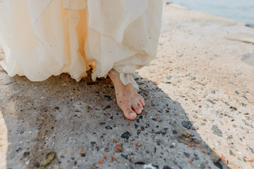 A young beautiful girl in a long milk-colored dress walks along the beach and pier against the background of the sea.