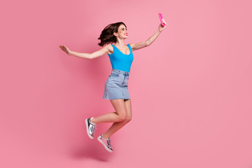 Full size profile photo of funny crazy lady jump high hold telephone making selfies speak talk skype wear casual blue tank-top denim short skirt shiny shoes isolated pink color background