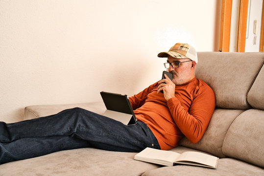 Adult with glasses, short beard and cap resting on the grey sofa of a modern and minimalist house. Connected, checking, using mobile devices, tablet, ipad, smartphone. Open book. White wall. 50´s. - Powered by Adobe