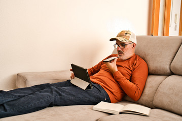 Adult with glasses, short beard and cap resting on the grey sofa of a modern and minimalist house. Connected, checking, using mobile devices, tablet, ipad, smartphone. Open book. White wall. 50´s.