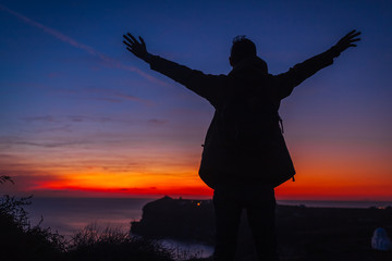 Fototapeta premium Man traveler silhouette hiking at sunset on Santorini island. Happy tourist raised arms feeling free, admiring landscape