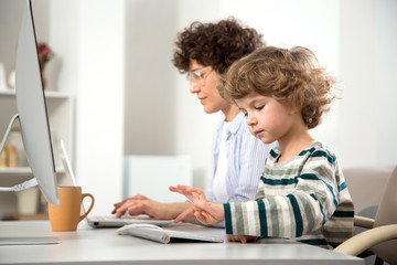 Business woman and her cute little son are using a computer and smiling while sitting in home office