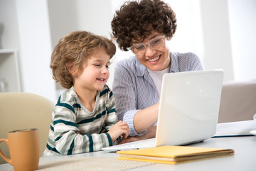Fototapeta premium Business woman and her cute little son are using a laptop and smiling while sitting in home office
