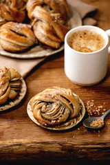 Traditional Swedish cardamom sweet buns Kanelbulle on cooling rack, ingredients in ceramic bowl above, cup of coffee on wooden table.