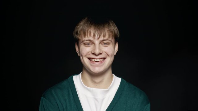 Close up of young man looking at camera and smiling against black background. Happy teenager with short brown hair.