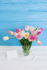 Tulips flowers in glass vase with gift card over blue wooden background for March 8, Women's Day, Birthday, Valentine's Day or Mother's day