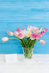 Tulips flowers in glass vase with gift card over blue wooden background for March 8, Women's Day, Birthday, Valentine's Day or Mother's day