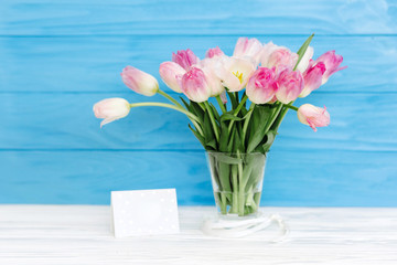 Tulips flowers in glass vase with gift card over blue wooden background for March 8, Women's Day, Birthday, Valentine's Day or Mother's day