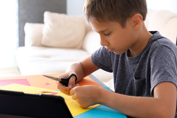 Naklejka premium Education, paper crafts for kids. Child cutting colored paper with scissors. Boy looking video tutorial and making paper craft project at home