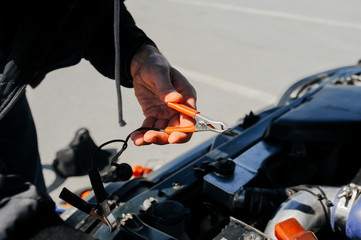 the hand connects the tongs red terminal to the battery of a car with an open hood and tuned parts of the car