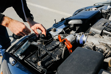 the hand connects the tongs red terminal to the battery of a car with an open hood and tuned parts...