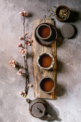 Tea drinking wabi sabi japanese style dark clay cups and teapot on wooden board with blooming cherry branches. Grey texture concrete background. Flat lay, space