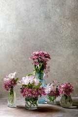 Group of glass bottles and vases with pink decorative flowers bouquets over grey texture background.