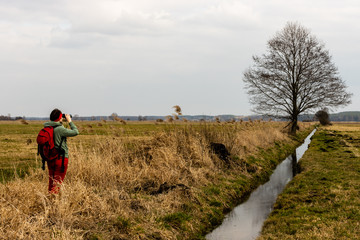 Obraz premium Female tourist person birdwatching in Grady Polewne Natura 2000 area. Woman in red using binoculars in grassland next to channel. Poland, Europe.