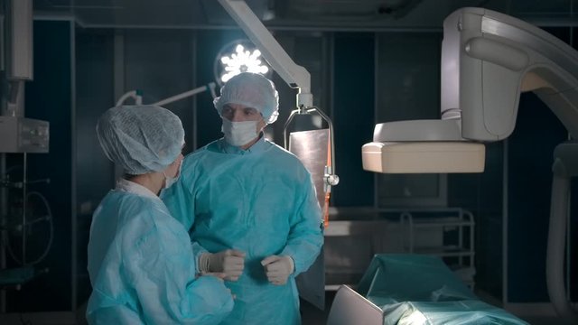 Two confident competent doctors discussing the plan of surgery. Adult male and young colleague assistant of surgery operation indoor of operating room. Slow motion.