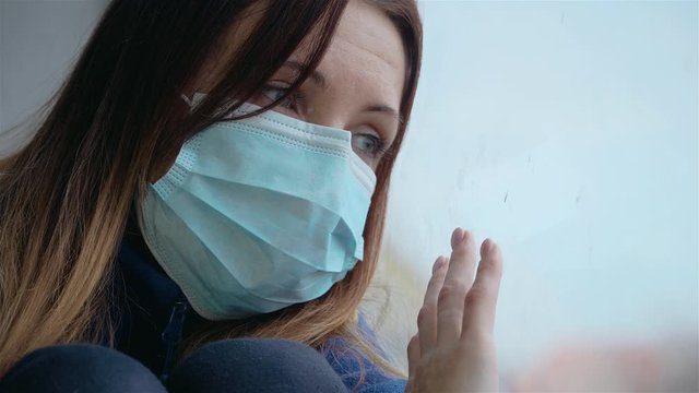 Woman at home looks out window in medical mask, quarantined due to spread of coronavirus. Stop COVID-19 infection