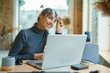Attractive pleased woman sitting at the desk with laptop looking away and smiling while working in coffee bar or coworking space. Concept remote work, freelance, using laptop computer or net-book.