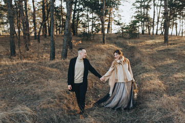 Fototapeta premium Stylish groom in a black cardigan and a beautiful bride in an expensive dress are walking in the woods in nature, holding hands. Wedding portrait of smiling newlyweds. Photography, concept.
