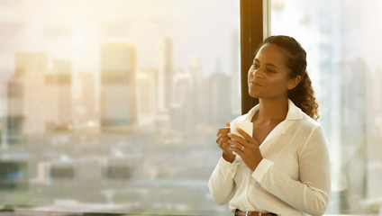 Attractive Young african american  business woman drinking  coffee while standing at  window in  office on cityscape background in morning light . happy black female enjoy tea relaxing on big city