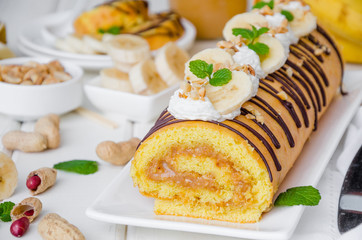 Biscuit or sponge cake roll with peanut butter cream, bananas and chocolate glaze on a white wooden background. Horizontal orientation. Close up.