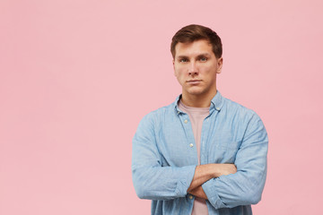 Waist up portrait of adult man standing with arms crossed and looking at camera while posing against pink background in studio, copy space