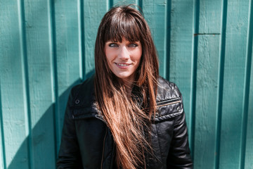 Fototapeta premium Portrait of a young woman in black coat leaning against a green wooden wall on a sunny afternoon.