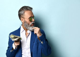 Senior man in white shirt, jacket and sunglasses. Holding a fan of hundred dollar bills, looking aside, posing on blue background