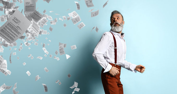 Elderly man in white shirt, brown pants and suspenders. He running from sheets of paper randomly located on photo, blue background