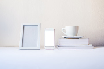 Fototapeta na wymiar White frame mock-up. Blank frame and mock up screen smartphone with white cup books by the white wall. Mock up frame concept. Minimalism concept. White concept