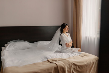 Beautiful happy bride sits on a sofa. Bride with elegant hairstyle and makeup. Wedding day. Beauty, fashion bride with long veil. Happiest bridal day.