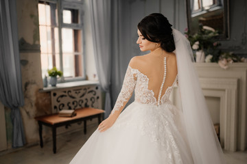 Fototapeta premium Elegant bride standing with her back to the camera holds her dress with her hands and looks profile. Full-length portrait of a bride in a vintage interior. Beautiful bride portrait.