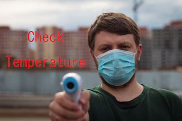 Young man in a green T-shirt in a medical mask holds a contactless thermometer on the street